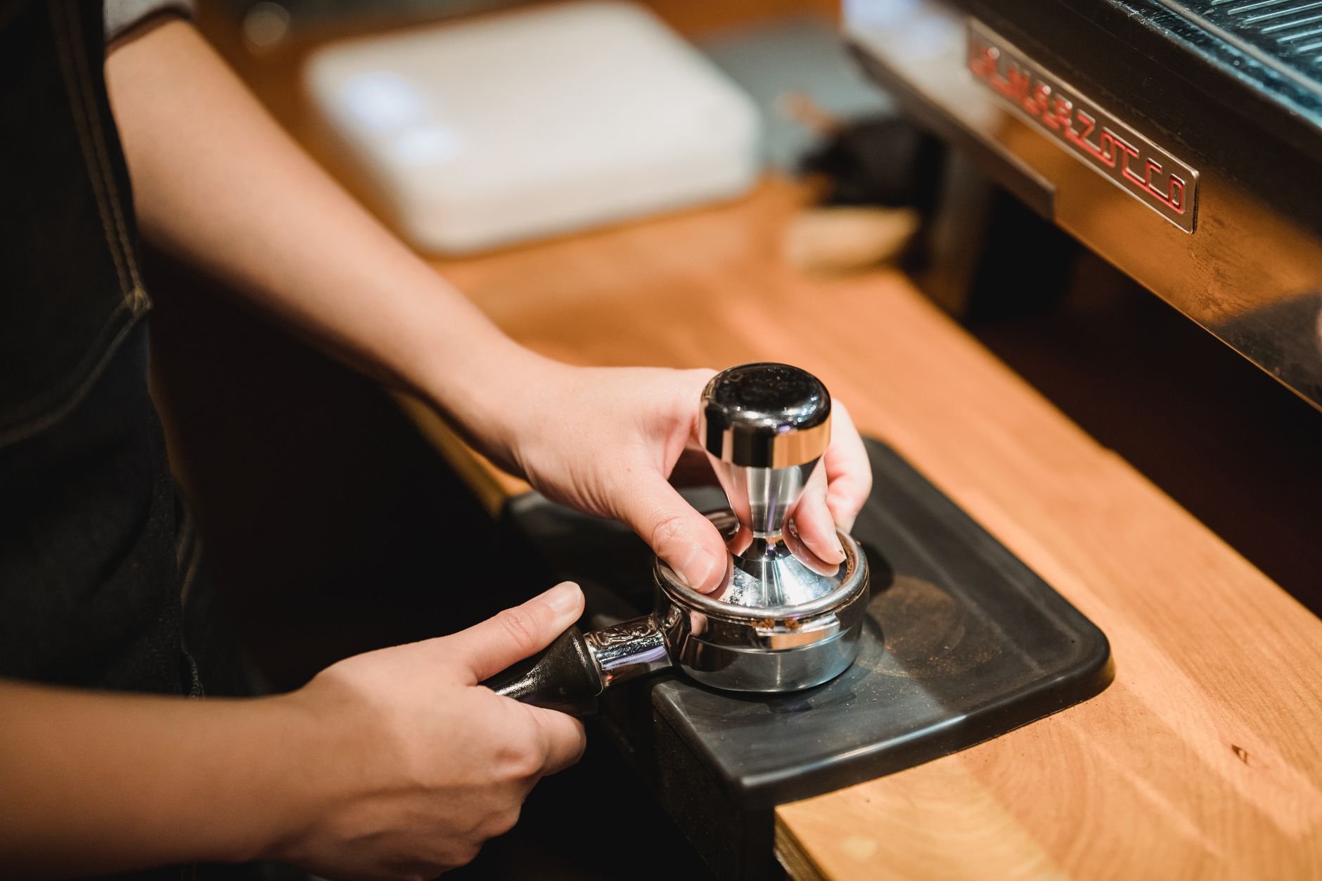 Close-up of barista hands tamping espresso grounds using a portafilter in a cozy café setting.